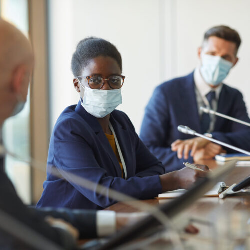 Portrait of African young businesswoman in protective mask looking at camera while sitting at meeting with her colleagues